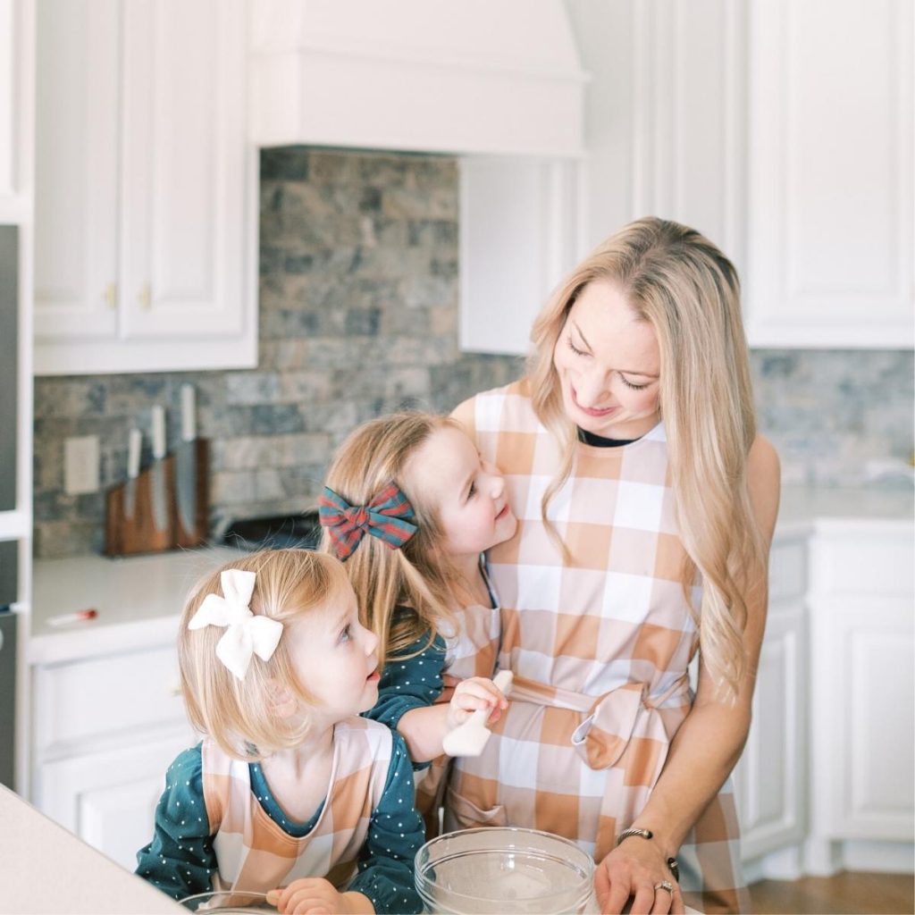 woman and 2 girls in brown checkered aprons in a white kitchen.