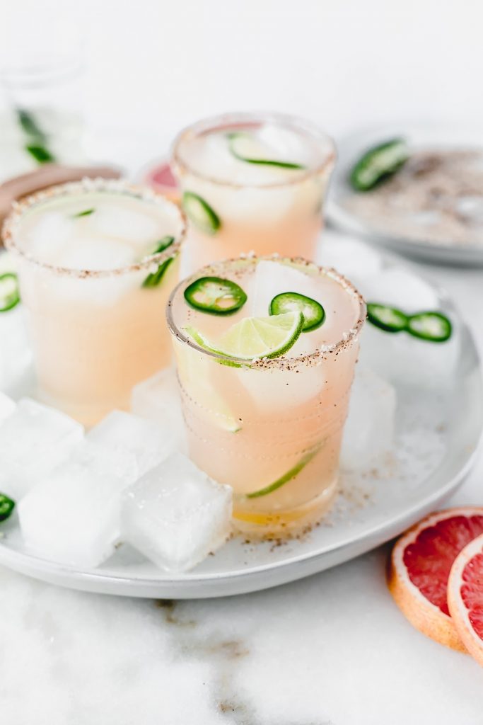 three mezcal paloma cocktails with lime and jalapeno slices in them on a plate with ice around them.