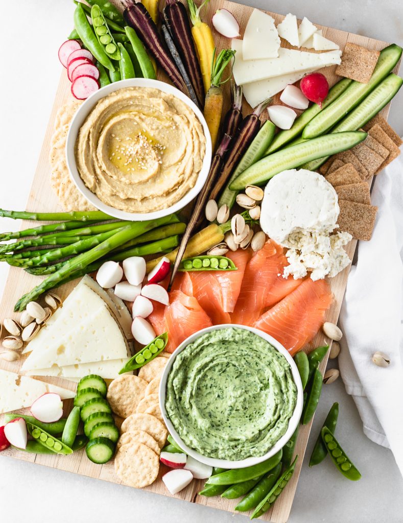 overhead view of a spring charcuterie board with hummus, green goddess dip, smoked salmon, cheeses, veggies and nuts.